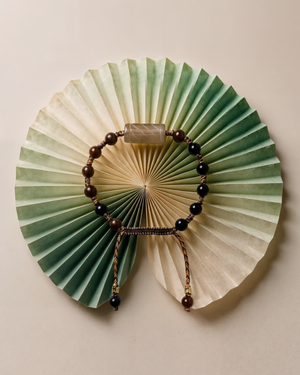 Bracelet of dark brown and black beads with a pale banded stone bead on braided cord, on a green paper fan.